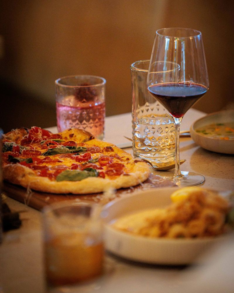 Pizza with toppings on a wooden board, next to a glass of red wine and a textured water glass on a dining table at dinner time.