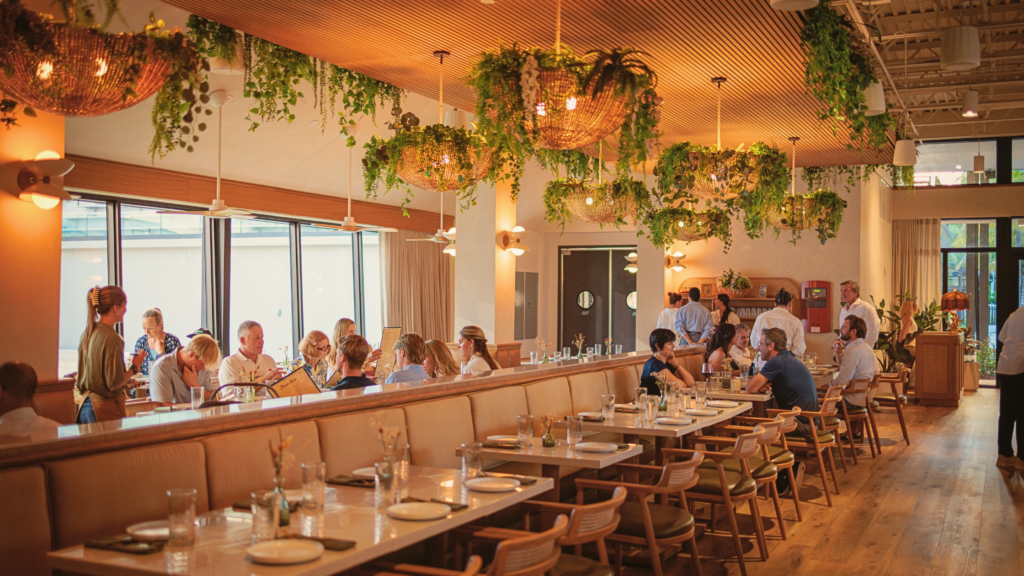 Elegant restaurant dining room at Florida Room in Fort Lauderdale with guests seated under warm lighting and greenery-filled ceiling fixtures